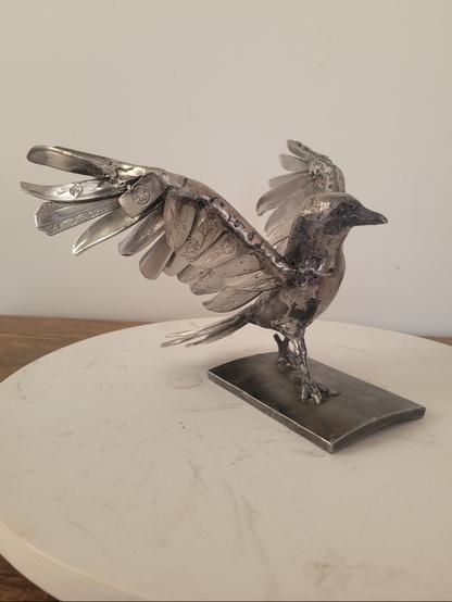 Sur la photo, une corneille en acier marche, les ailes déployées comme pour s'envoler mais de manière très décidée.