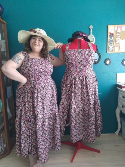 Posing with the Rosie dress by mercurea brand, mercurea wears it with the ties laced at the shoulders and the mannequin has them tied behind the neck. This dress is a rose printed cotton fabric with Bougainville coloured velveteen lace decorations