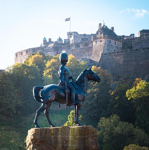 EN: The completely black statue shows a cavalry rider in typical uniform looking into the distance from his horse. Rifles hang from the saddle on either side. The sculpture stands on a memorial stone.
Castle Hill rises up in the background with its trees already slightly coloured in autumn and the north-eastern buildings of Edinburg Castle.

DE: Die komplett schwarze Statue zeigt einen Kavalleriereiter in typischer Uniform, der von seinem Pferd in die Ferne schaut. Gewehre hängen zu beiden Seiten vom Sattel. Die Skulptur steht auf einem Gedankestein.
Im Hintergrund erhebt sich Castle Hill mit schon leicht herbstlich gefärbten Bäumen sowie den nordöstlichen Gebäuden von Edinburg Castle.
