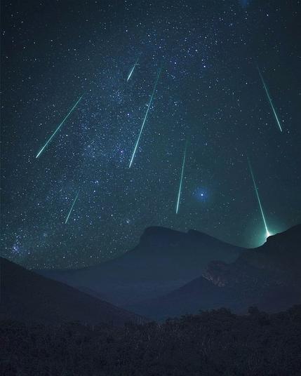 A breath-taking view of a meteor shower with bright green streaks of light falling across a star-filled night sky above the silhouetted peaks of the Grampians mountains.