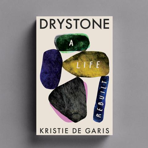 A colour image of a book cover. There are five large stones coloured green, purple, ochre, grey and blue stacked in a pleasing way. On the green, ochre and blue stones are the words 'A Life rebuilt' The title is in black at the top of the image and says 'Drystone'. The authors name, at the bottom of the image, is Kristie De Garis.
