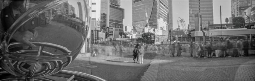 Panoramic long exposure black and white pinhole photo of Shibuya Crossing with motion blur and reflection of camera and photographer in sliver globe on left foreground.