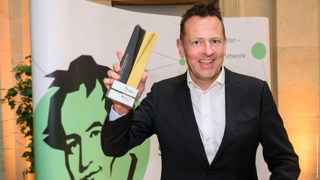 Foto: Person im schwarzen Anzug, Andreas Winter,  hält einen schwarz-goldenen Preis vor einem Hintergrund mit grün-schwarzem Gesichtsporträt.