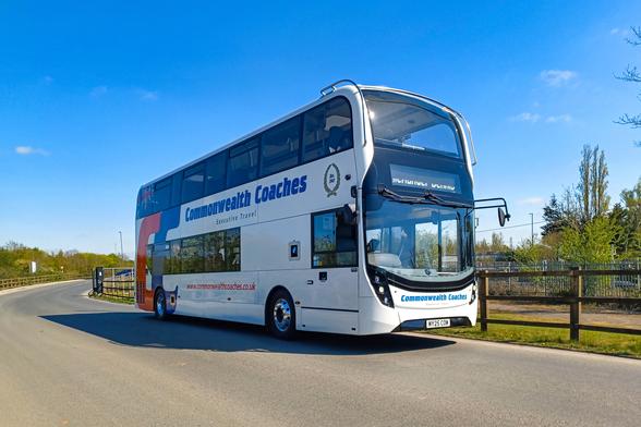 The image shows a double-decker bus branded "Commonwealth Coaches" parked at the roadside. The bus is mainly white with blue and red detailing, and large windows on both decks. The road curves gently into the background, bordered by a wooden fence and greenery, with trees visible in the distance. The sky is clear and blue, indicating a sunny day. The front of the bus features a large windscreen and side mirrors.