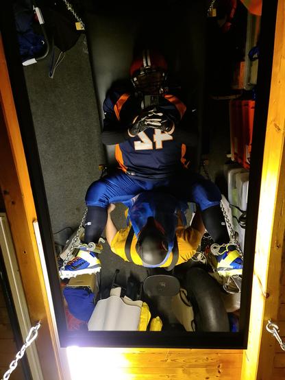 picture taken via ceiling mirror: person wearing full American football gear lying on a fuckboard, person in orange t-shirt and pup hood standing in front of him