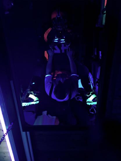 picture taken via ceiling mirror: person wearing full American football gear lying on a fuckboard, person in orange t-shirt and pup hood standing in front of him, dark scene lit by UV/blacklight