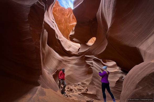 A slot canyon with smooth, curving walls in deep reddish-brown hues. Light from the open sky above filters down, illuminating the intricate patterns formed by erosion. Two people stand inside—one capturing a photo of the other—framing the natural beauty of the canyon. The scene highlights the dramatic contrast between shadowed rock formations and bright sunlight above, emphasizing the canyon's depth and textures.