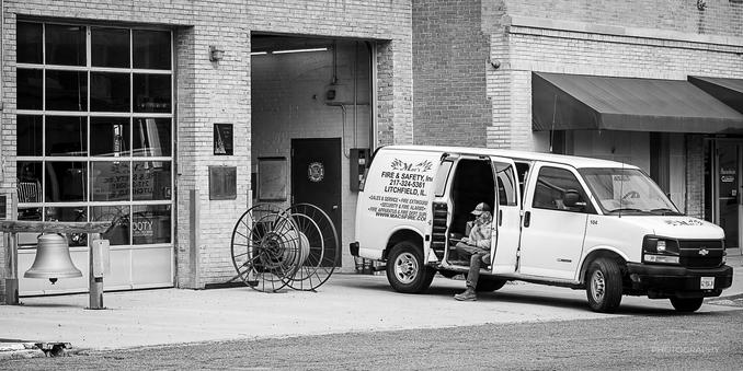 A man sits in the open side doors of a van from Mac's Fire & Safety in the front of a fire department.