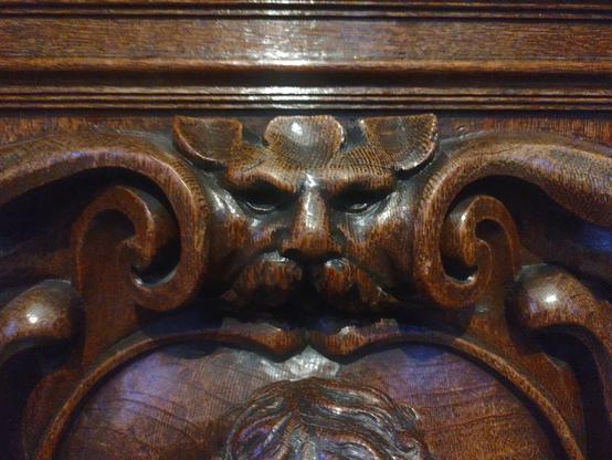 The image shows a detailed wooden carving of a man's face with beard on what appears to be antique furniture, likely a cabinet or chest. The carving features a rich, polished dark wood with detailed texturing showing the dog's features including its muzzle and ears. The wood has a glossy finish that reflects light, and the carving is part of a larger decorative panel with scrollwork and other ornamental elements surrounding it.