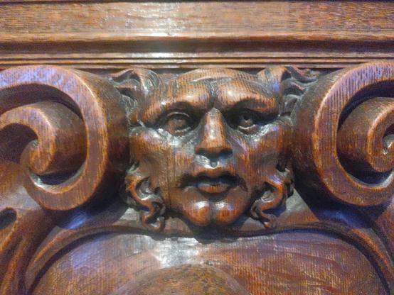 The image shows a detailed wooden carving of a face on what appears to be antique furniture, likely a cabinet or chest. The carving features a rich, polished dark wood with detailed texturing showing the dog's features including its muzzle and ears. The wood has a glossy finish that reflects light, and the carving is part of a larger decorative panel with scrollwork and other ornamental elements surrounding it.