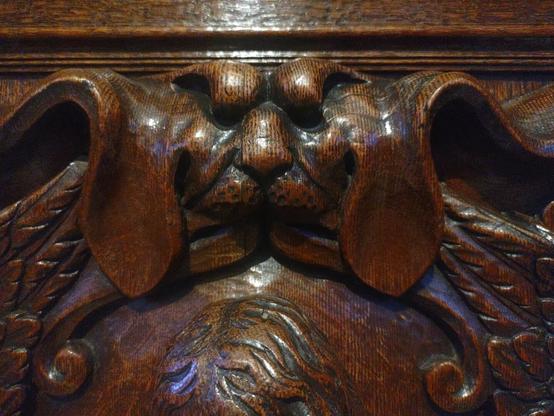The image shows a detailed wooden carving of a dog's face with prominent ears on what appears to be antique furniture, likely a cabinet or chest. The carving features a rich, polished dark wood with detailed texturing showing the dog's features including its muzzle and ears. The wood has a glossy finish that reflects light, and the carving is part of a larger decorative panel with scrollwork and other ornamental elements surrounding it.