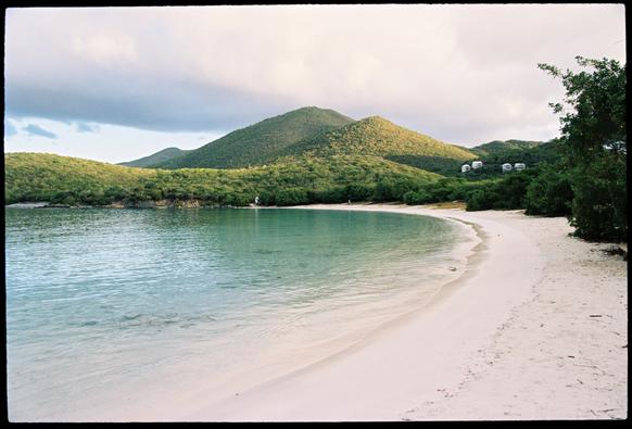 A film photo of a crescent-shaped sand beach taken from near the water’s edge. The water is a bright emerald-turquoise and there are densely vegetated mountains in the background. Early morning sunlight touches parts of the mountains, giving them depth and texture.