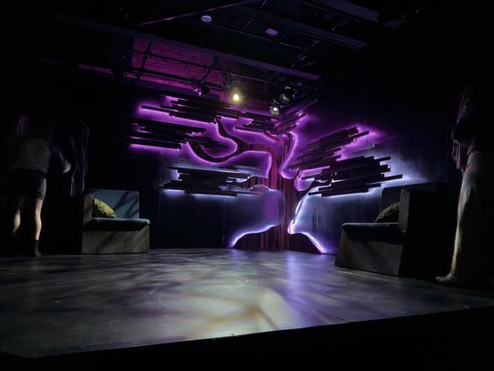 A mesmerizing stage design featuring an ethereal tree installation that transforms the dark performance space into a dreamlike environment. The centerpiece is a stylized tree created with rich wooden elements – a flowing, organic silhouette with curved branches that seem to dance across the wall. Surrounding this central form are numerous wooden slats arranged in horizontal layers that extend outward like an artistic interpretation of foliage or energy radiating from the tree.
The entire installation is dramatically backlit with vibrant turquoise LED lights that create a stunning contrast against the warm wood tones. The aqua blue glow seeps through gaps between each wooden plank and traces the undulating curves of the tree's form, casting an otherworldly aura throughout the darkened space. The lighting technique creates depth and dimension, making the wooden elements appear to float against the black walls.
