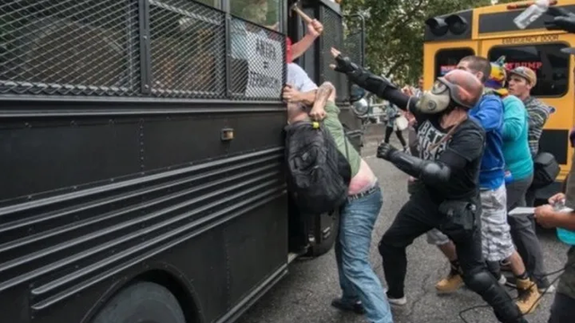 June 2019 neo-nazi attack in Portland, one nazi is trying to swing a hammer at counter protesters, Alexander Dial clad in black, a helmet, gas mask and protective gear is reaching up to block the hammer before it can be swung from a black bus.