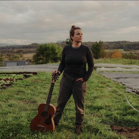 An AFAB person with long brown hair tied in a bun stands in a vegetable patch. They wear a grey sweater and dark brown pants. They are holding a guitar and look off to the right.