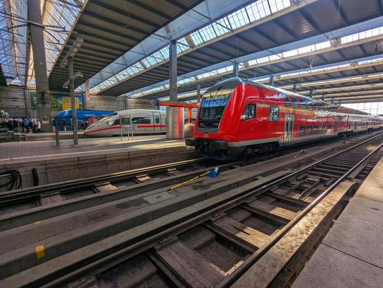 The image shows the Munich Hauptbahnhof, featuring a red DB Regio train on the tracks. Another train, possibly a Deutsche Bahn ICE, is visible in the background. The station has a large, open structure with a glass and metal roof.