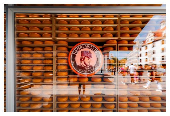 Eine vitrine mit zahlreichen großen, runden Käselaiben in leuchtendem Orange ist das zentrale Objekt. Im Vordergrund befindet sich ein rundes Emblem mit dem Schriftzug „HENRI WILLIG SINCE 1974“. Im Hintergrund sind Passanten zu sehen, die eine belebte Straße durchqueren, und historische Gebäude spiegeln sich in der Glasfront der Vitrine. Die Szene vermittelt ein geschäftiges, einladendes Ambiente.