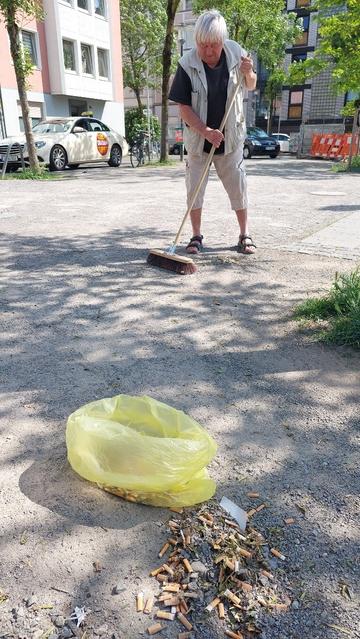 Eine Person kehrt Zigaretten auf dem Boden zusammen. Im Vordergrund liegt eine Mülltüte neben einem Haufen Zigarettenkippen.
