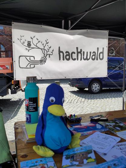 Im Vordergrund sitzt ein genähter Pinguin zwischen dem Infomaterial und Pixi-Büchern, dahinter steht präsent eine Trinkflasche mit dem Sticker "Das Internet zerstört dein Leben.". Im Hintergrund ist an einem schwarzen Sonnenschutz mit einer Kite-Leine ein wunderschönes weißes Banner mit dem hackwald-Logo aufgehangen. Ganz hinten stehen Autos auf gepflastertem Grund,