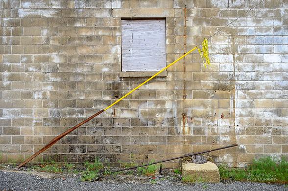 A concrete block building with a boarded up window.  In front are a wire pole support wrapped in yellow tape, a small metal pole, and a concrete foundation of a broken off wooden telephone pole.