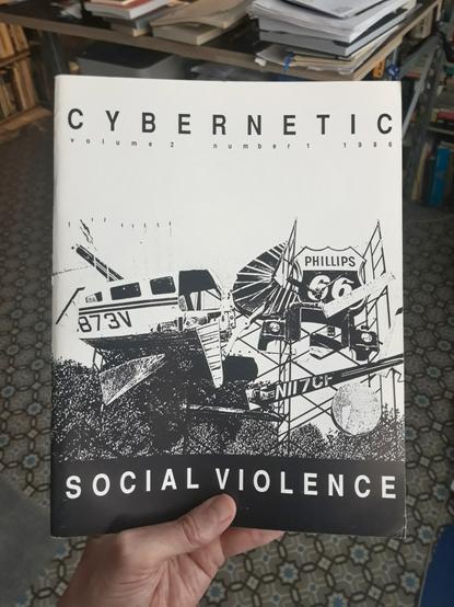 A hand holds a copy of the 'Cybernetic' magazine, Volume 2, Number 1, 1986, entitled 'Social Violence'. The black and white cover shows what looks like a photocopied photograph of a helicopter that crashed into a publicity sign of some sort.