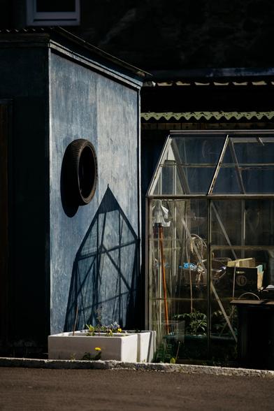A blue-painted shed wall with a mounted tyre casting a strong geometric shadow. Beside it, a weathered glass greenhouse filled with tools and baskets, bathed in late-afternoon sunlight