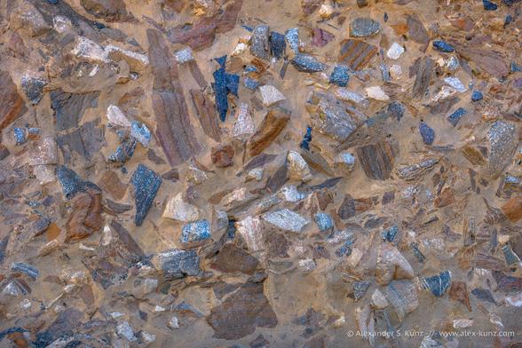 Close-up photo of a rock conglomerate with colorful pieces stuck in a beige-brown matrix, like a solidified landslide.