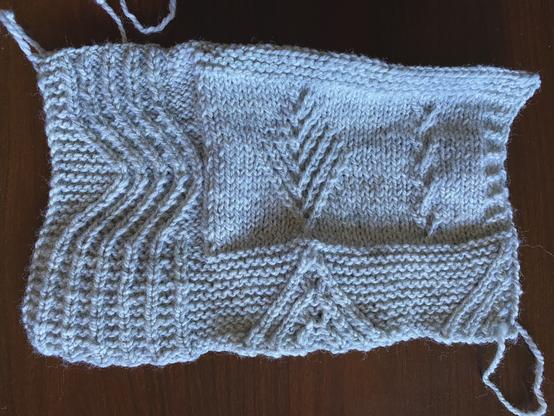 Hand knit swatch in a light bluish grey, with twisted stitch patterns of waves on the left, mountains against garter stitch on the bottom, sort of conifer trees in the middle, and something that when I play around with the placement will hopefully evoke grasslands. The swatch is unblocked, so the stitches are a bit wobbly and the top garter border is curling up a bit.