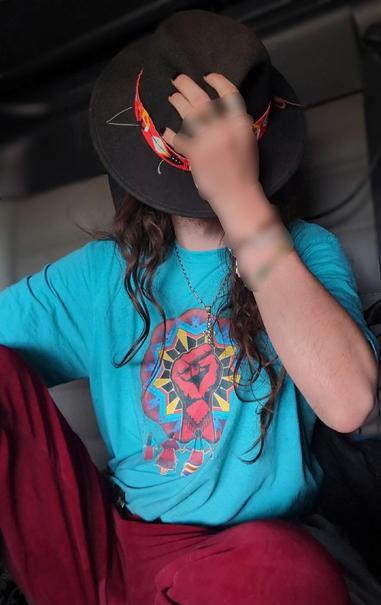 Me wearing red pants and torquoise tshirt with a print  of a red fist above a semi-circle of NDN women holding hands. A black hat with a red beaded band covers his face.