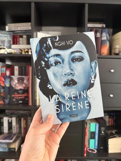 Livre pris en photo devant une bibliothèque. La couverture de La reine sirène montre un portrait de femme asiatique dans un ton bleuté/noir et blanc, maquillée, chignon et boucle d’oreilles, expulsant de la fumée de sa bouche entrouverte.