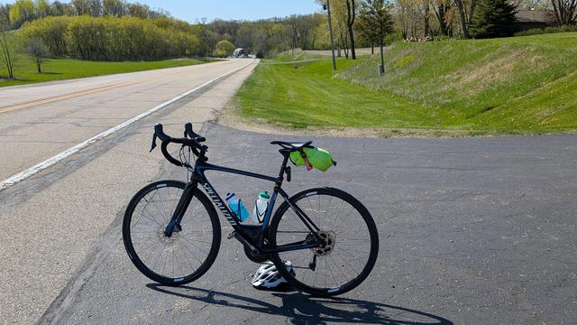 Blue and black Roubaix bicycle parked in an asphalt driveway on the right of Olmsted County 3 which heads downhill and curves to the left. On either side are green lawns and trees with new green leaves. At the bottom of the hill is a red barn,