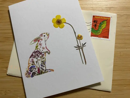 A greeting card, white, a bunny made up of spring flowers standing on its back feet stretching towards a tall yellow flower.
Underneeth a cream coloured envelop with a red stamp showing a green snake and Chinese writing.
Both a laying on a wooden surface.