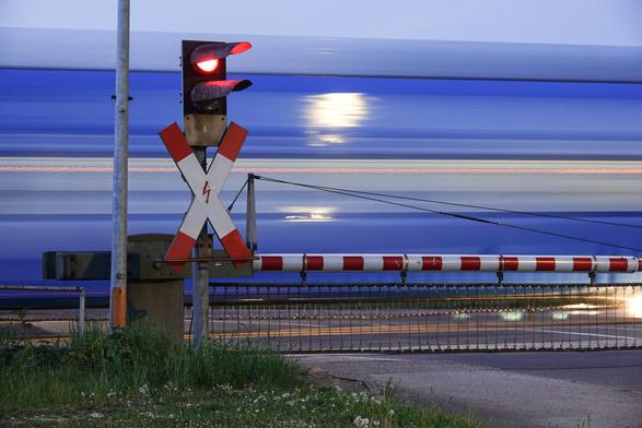 Es ist abends. Lichtzeichen mit rot leuchtenden Licht sowie geschlossener Schranke vor einem durchfahrenden Zug in blauer Farbe.