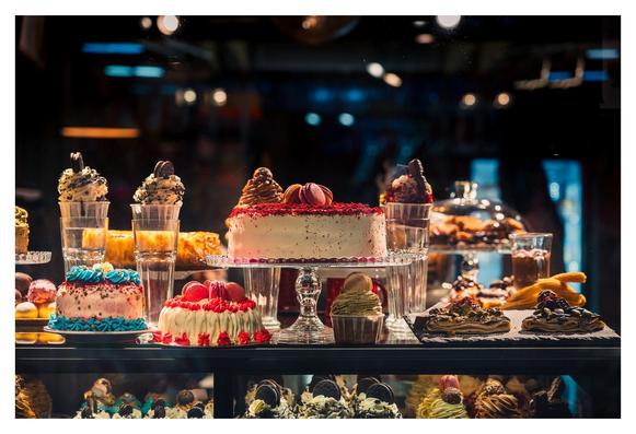 Farbenfrohe Torten und Süßigkeiten stehen kunstvoll hinter einer Glasvitrine. Ein großer, dekorierter Kuchen mit roten und rosa Akzenten zieht die Blicke an, während verschiedene kleine Törtchen und Eistüten angrenzen. Die Umgebung strahlt einladendes Licht aus, das die Leckereien hervorhebt. Gläser mit Desserts und hellem Obst ergänzen die Auswahl, die an eine Konditorei erinnert und zum Genießen einlädt.