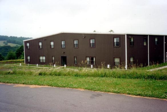 A long brown warehouse front, with windows on the first and second story, sitting in a green field with a gray sky and distant green hills. No cars, no people, definitely in the middle of nowhere, Pennsylvania.