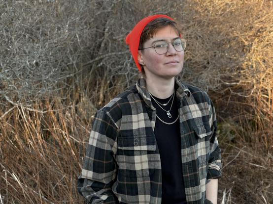 Waist-up color photo of writer Anneke Schwob, a fair-skinned person with brown hair worn short with bangs peeking out from beneath a red beanie. They also wear wire-rimmed glasses, a green-brown-and-white plaid flannel worn open over a black crew-neck tee,  three chunky silver necklaces extending from Anneke’s collarbone height to height of estimated mid-sternum, and multiple earlobe piercings. They stand in a field of dry, yellowing grasses, their elbows bent as if their hands are in the pockets of their offscreen trousers. They are turned slightly away from the camera, looking into the lens with serious, steady expression that may hold a slight smile.