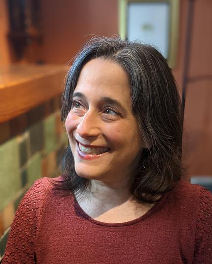 Head-and-shoulders color photo of writer Jennifer Hudak, a fair-skinned woman with straight, gray-threaded brown hair worn at shoulder length and without bangs. She wears a brownish-red top with crochet sleeves and poses before a blurred interior background that includes tile wall and light wood. Her shoulders are square to the camera, but she looks up and to one side, smiling warmly.