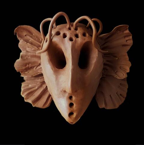 The image shows a plasticine sculture with an elongated, triangular face shape against a black background. It has hollow eye sockets and several small holes across the upper portion of the face. The mask features decorative elements including curling tendrils or loops extending from the top, and large fan-like or ruffled structures on both sides resembling ears or wings. The piece has a warm reddish-brown color typical of natural clay. The artistic mask appears to be inspired by ceremonial or ritual designs, with its distinctive stylized features and textural elements creating a dramatic, somewhat otherworldly appearance.