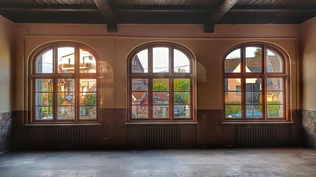 Opuszczone wnętrze dworca. Widok na trzy łukowato zakończone okna, za nimi krajobraz miejscowości.