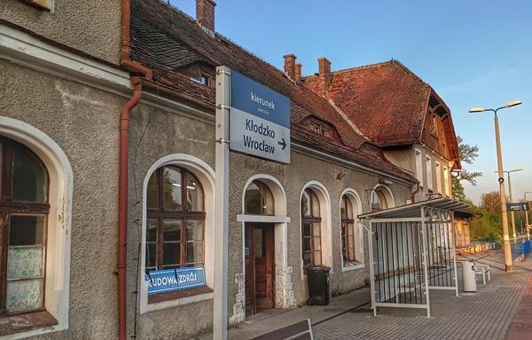 Dworzec w Kudowie-Zdroju od strony peronu. Tablica z nazwą stacji popękana i położona na ramie okiennej. Na peronie nowa wiata i oznaczenie kierunku trasy do Kłodzka i Wrocławia.