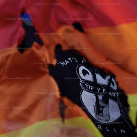 A burnt rainbow flag (the red, orange and yellow stripes are visible) with holes burnt into it. Through the biggest one a black hoodie of Tipsy Bar Berlin can be seen. The logo is an illustration of a blindfolded bear.
