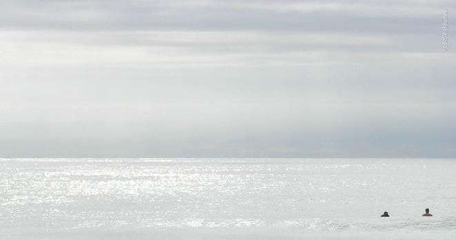 A calm silver sea meets a silver clouded horizon in an almost monotone, abstract tonal image. Near the lower right corner two contrasting black dots, the dark heads and shoulders of two distant swimmers, provide scale and contrast