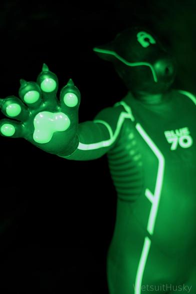 Person wearing a black neoprene wetsuit with a BLUE70 logo on the chest, lit dramatically with green lighting. They are also wearing a black Furrjoi Drone Mask resembling a canine head and extending a gloved paw toward the camera. The glove features glowing green paw pads, creating a bold, futuristic, and kink-inspired visual.