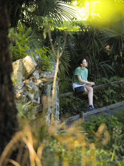 La photographie représente une personne assise sur une balustrade en bois dans une zone verdoyante. De nombreuses plantes et fougères l'entourent. Une petite chute d'eau est visible à gauche et l'éclairage suggère un cadre paisible et ensoleillé.