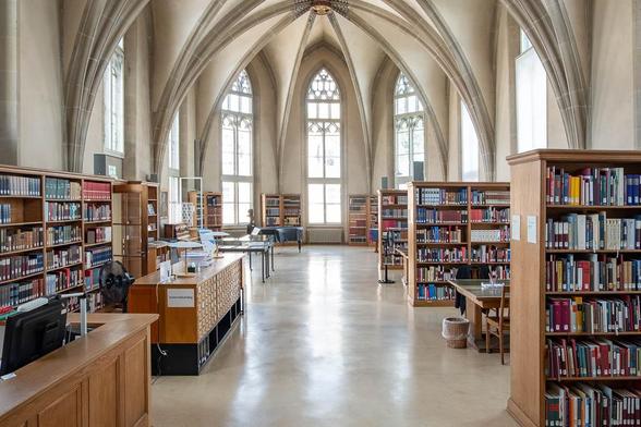 A library room in a former church with tall Gothic windows, high vaulted ceilings, wooden bookshelves filled with books, desks and card catalogues.