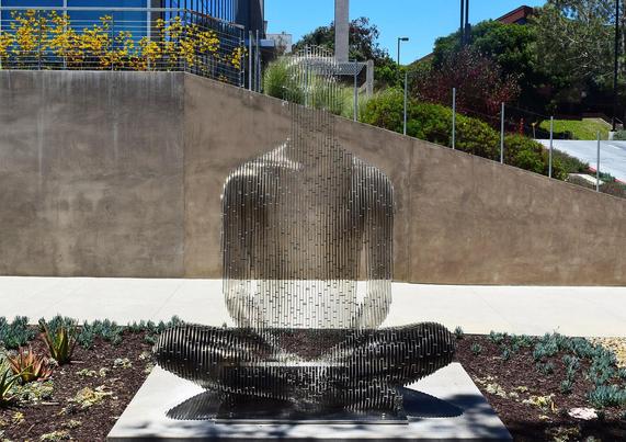 A minimalist sculpture of a seated figure, crafted from vertical metal rods, reflecting light amidst landscaped surroundings.