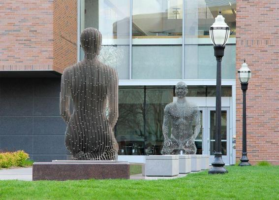 Two abstract metal sculptures of human figures stand outdoors, framed by a modern building and street lamps, surrounded by green grass.