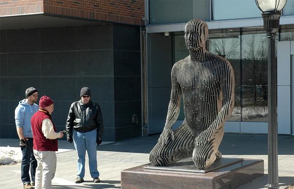 A sleek, modern metal sculpture of a kneeling figure sits on a pedestal outside a building, surrounded by three people.