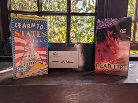 three tapes on a mantel. glowing green window behind. 

- learning the usa capital cities 
- DEAD FIRE
- something labeled "fbc on air promotions"