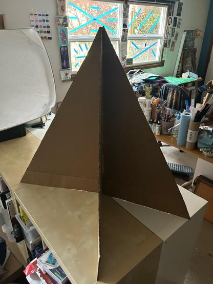 A tall tree shape made of cardboard sitting on a work surface. Windows with blue tape (to deter birds) are in the background as is a desk with lots of art supplies handy. A light tent is odd to the left side.